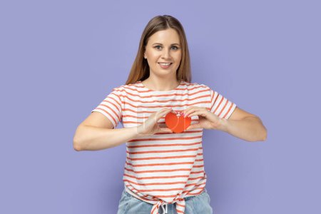 Photo for Portrait of smiling adorable blond woman wearing striped T-shirt holding little red heart in front her chest, expressing romantic feeling and love. Indoor studio shot isolated on purple background. - Royalty Free Image