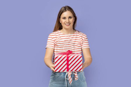 Photo for Portrait of satisfied attractive festive blond woman wearing striped T-shirt giving present box, congratulating her friend with holiday. Indoor studio shot isolated on purple background. - Royalty Free Image