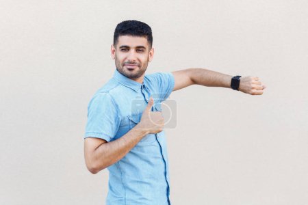 Photo for Portrait of positive attractive calm man wearing denim shirt standing showing his smart watch and thumb up, like gesture, recommend. Indoor studio shot isolated on gray background. - Royalty Free Image