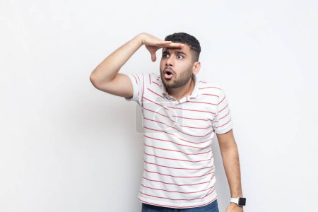 Photo for Portrait of amazed concentrated impressed bearded man wearing striped t-shirt standing with hand near forehead, looking far with open mouth. Indoor studio shot isolated on gray background. - Royalty Free Image