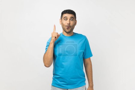 Photo for Portrait of excited amazed clever smart unshaven man wearing blue T- shirt standing with finger up, having idea, looking at camera with surprised face. Indoor studio shot isolated on gray background. - Royalty Free Image