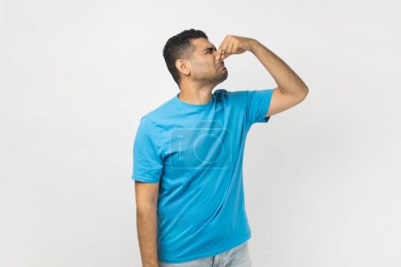 Photo for Portrait of disgusted handsome unshaven man wearing blue T- shirt standing and pinching nose, smelling awful odor, turning his face. Indoor studio shot isolated on gray background. - Royalty Free Image
