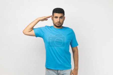 Photo for Portrait of serious strict handsome unshaven man wearing blue T- shirt standing with hand near, saying yes sir, being ready to do command. Indoor studio shot isolated on gray background. - Royalty Free Image