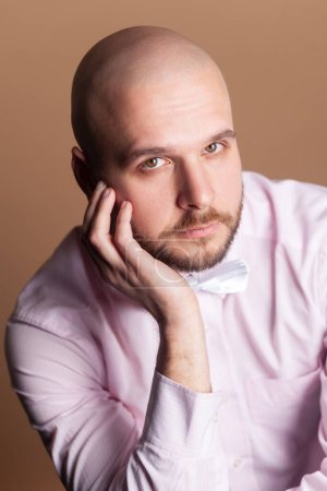 Photo for Portrait of calm serious attractive bald bearded man sitting keeps hand on his face, looking at camera, wearing light pink shirt and bow tie. Indoor studio shot isolated on brown background. - Royalty Free Image