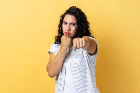 Photo for Portrait of aggressive woman with dark wavy hair holding clenched fists up, fighting with abuser, defence from domestic violence. Indoor studio shot isolated on yellow background. - Royalty Free Image