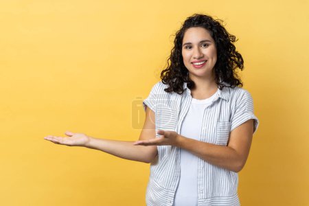 Photo for Portrait of smiling satisfied woman with dark wavy hair showing welcome gesture and empty space on wall for your best advertising. Indoor studio shot isolated on yellow background. - Royalty Free Image