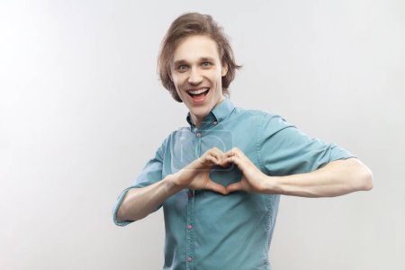 Photo for Portrait of smiling cheerful young man showing heart shape with fingers, expressing love, affection, looking at camera, wearing blue shirt. Indoor studio shot isolated on gray background. - Royalty Free Image