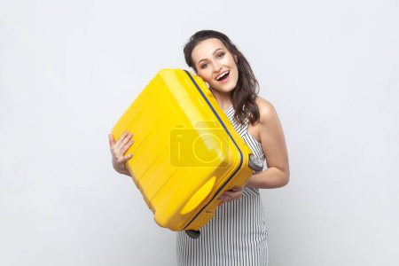 Téléchargez les photos : Portrait de femme brune positive souriante et joyeuse debout avec une valise jaune, se réjouit de son futur voyage, portant une robe rayée. Studio intérieur tourné isolé sur fond gris. - en image libre de droit