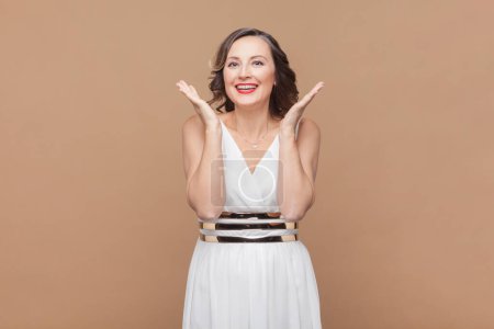 Photo for Portrait of joyful middle aged woman with wavy hair keeps hands raised looks positively at camera and laughing, wearing white dress. Indoor studio shot isolated on light brown background. - Royalty Free Image