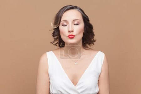 Photo for Passionate tender woman with wavy hair sends air kiss with folded lips, stands with eyes closed, looks cute and sensual, wearing white dress. Indoor studio shot isolated on light brown background. - Royalty Free Image