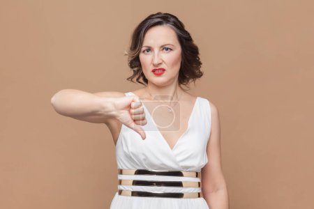 Photo for Discontent woman with wavy hair shows disapproval sign, keeps thumb down, expresses dislike, frowns face in discontent, wearing white dress. Indoor studio shot isolated on light brown background. - Royalty Free Image
