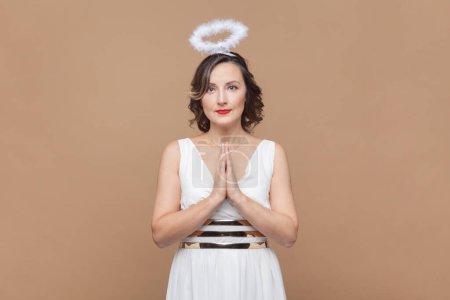 Photo for Portrait of hopeful middle aged woman with wavy hair and nimb over head, praying, pleading about something, wearing white dress. Indoor studio shot isolated on light brown background. - Royalty Free Image