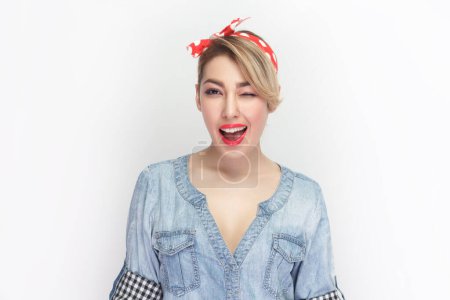 Photo for Portrait of positive happy charming blonde woman wearing blue denim shirt and red headband standing smiling joyfully and winking eye. Indoor studio shot isolated on gray background. - Royalty Free Image