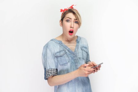 Photo for Portrait of shocked surprised blonde woman wearing blue denim shirt and red headband standing holding mobile phone, looking at camera with big eyes. Indoor studio shot isolated on gray background. - Royalty Free Image