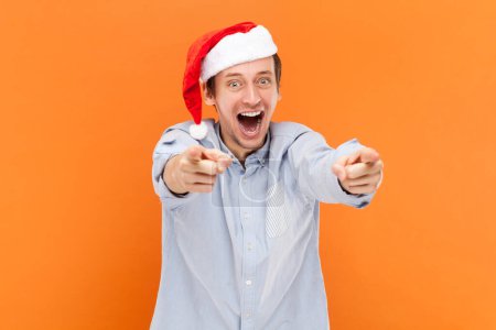 Téléchargez les photos : Portrait d'un beau jeune homme adulte émerveillé pointant du doigt la caméra, vous choisissant, portant une chemise bleu clair et un chapeau du Père Noël. Studio intérieur tourné isolé sur fond orange. - en image libre de droit
