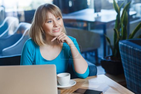 Photo for Portrait of satisfied delighted young woman with blonde hair in blue shirt working on laptop, having rest, drinking coffee or tea, looking away, dreaming with hand on chin. Indoor shot in cafe. - Royalty Free Image