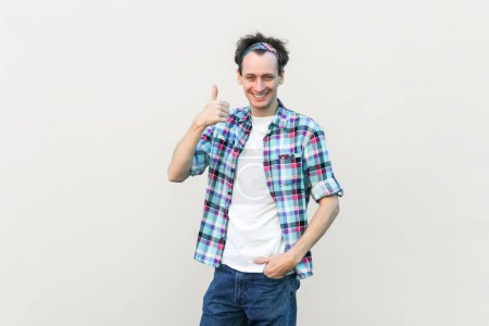 Photo for Portrait of man in casual blue checkered shirt and headband approves incredible promo, keeps thumbs up likes and agrees being satisfied with something. Indoor studio shot isolated on gray background. - Royalty Free Image