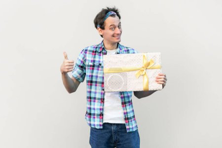 Photo for Portrait of handsome satisfied handsome man holding present box, celebrating holiday, showing thumb up, wearing blue checkered shirt and headband. Indoor studio shot isolated on gray background. - Royalty Free Image