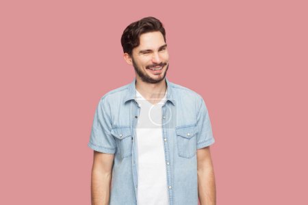 Photo for Portrait of playful positive bearded man in blue casual style shirt standing blinking his eyes with pleasure, having happy expression. Indoor studio shot isolated on pink background. - Royalty Free Image
