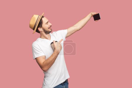 Foto de Retrato del bloguero barbudo encantado en camiseta blanca y sombrero de pie con teléfono inteligente en las manos y transmitiendo transmisión en vivo, bebiendo café. Estudio interior plano aislado sobre fondo rosa. - Imagen libre de derechos