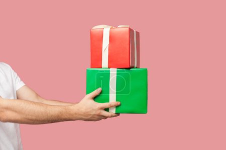 Téléchargez les photos : Portrait d'homme inconnu en T-shirt blanc debout montrant des boîtes cadeaux rouges et vertes, cadeaux pour les vacances. Studio intérieur tourné isolé sur fond rose. - en image libre de droit