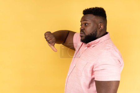 Photo for Side view portrait of sad upset bearded man wearing pink shirt showing thumb down, expressing bad negative emotions, dislike, frowning face. Indoor studio shot isolated on yellow background. - Royalty Free Image