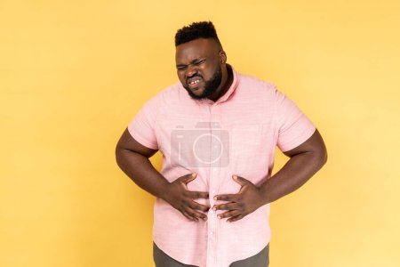 Photo for Health problems. Portrait of unhappy unhealthy sick man wearing pink shirt standing, holding her belly with hands, stomach cramps. Indoor studio shot isolated on yellow background. - Royalty Free Image