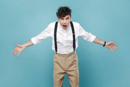 Photo for Portrait of angry aggressive man wearing white shirt and suspender standing and screaming with open mouth and raised arms. Indoor studio shot isolated on blue background. - Royalty Free Image