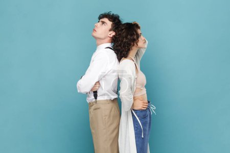 Photo for Portrait of couple or friends standing back to back to each other looking up with pensive thoughtful expression thinking about problems in relationship. Indoor studio shot isolated on blue background. - Royalty Free Image