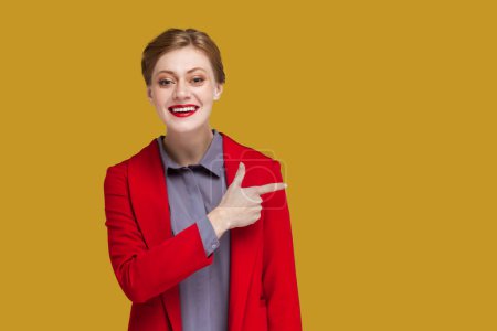 Téléchargez les photos : Portrait d'une femme souriante heureuse et positive avec des lèvres rouges pointant du doigt vers l'espace de copie pour la publicité, portant une veste rouge. Studio intérieur tourné isolé sur fond jaune. - en image libre de droit