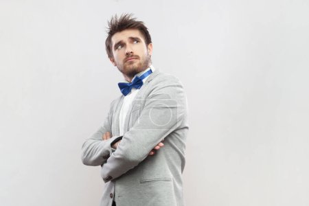 Photo for Portrait of confident pensive thoughtful bearded man standing folded hands, looking away, planing and pondering, wearing grey suit and blue bow tie. Indoor studio shot isolated on gray background. - Royalty Free Image