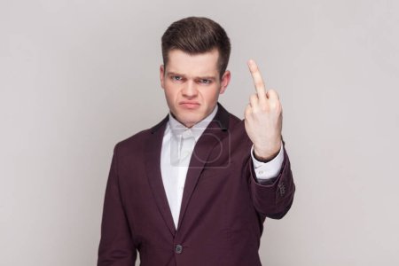 Foto de Portrait of man shows fuck off sign being rude and looks dissatisfied at camera, expresses bad attitude to someone, wearing violet suit and white shirt. Indoor studio shot isolated on grey background. - Imagen libre de derechos