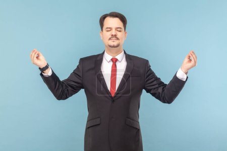 Photo for Portrait of relaxed man with mustache standing feels peaceful, practicing yoga, relaxing, wearing black suit with red tie. Indoor studio shot isolated on light blue background. - Royalty Free Image