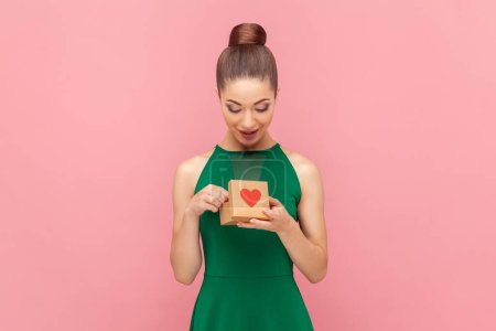 Téléchargez les photos : Portrait de femme joyeuse optimiste positive debout avec une petite boîte cadeau, regardant à l'intérieur, admire son cadeau, portant une robe verte. Studio intérieur tourné isolé sur fond rose. - en image libre de droit