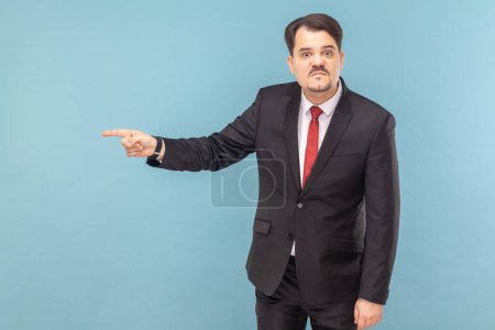 Photo for Portrait of angry annoyed serious man with mustache standing standing pointing at exit, saying get out, wearing black suit with red tie. Indoor studio shot isolated on light blue background. - Royalty Free Image