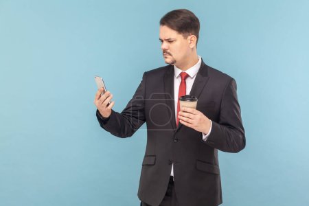 Photo for Portrait of serious concentrated man with mustache standing using mobile phone and drinking takeaway coffee, wearing black suit with red tie. Indoor studio shot isolated on light blue background. - Royalty Free Image