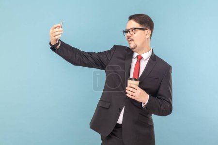 Photo for Portrait of funny man with mobile phone and making selfie or broadcasting livestream while drinking coffee, wearing black suit with red tie. Indoor studio shot isolated on light blue background. - Royalty Free Image