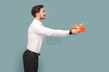 Photo for Side view portrait of friendly positive optimistic man with mustache giving red present box, congratulating with holiday, wearing Indoor studio shot isolated on light blue background. - Royalty Free Image