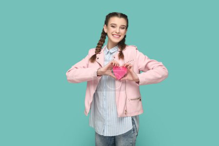 Téléchargez les photos : Portrait d'adolescente ravissante et souriante avec des tresses portant une veste rose tenant un petit cœur rose, regardant la caméra, exprimant de l'amour. Studio intérieur tourné isolé sur fond vert. - en image libre de droit