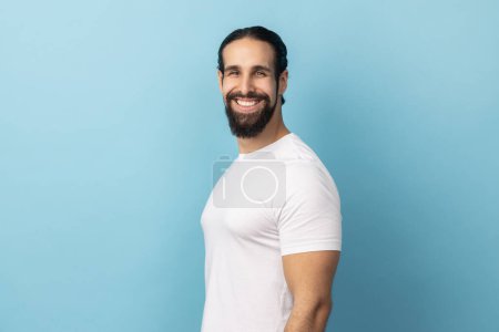 Photo for Side view of bearded handsome man wearing white T-shirt standing looking at camera with satisfied face and smiling, expressing happiness. Indoor studio shot isolated on blue background. - Royalty Free Image