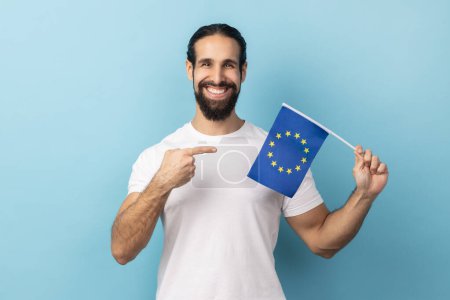 Photo for Portrait of man with beard wearing white T-shirt smiling broadly and pointing flag of European Union, symbol of Europe, EU association and community. Indoor studio shot isolated on blue background. - Royalty Free Image