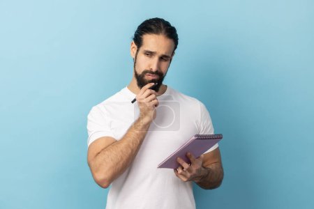 Photo for Portrait of thoughtful man with beard wearing white T-shirt holding paper notebook, having thoughtful facial expression, planning. Indoor studio shot isolated on blue background. - Royalty Free Image