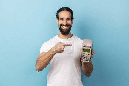 Photo for Portrait of joyful man with beard wearing white T-shirt pointing finger at pos terminal, suggesting you to use contactless payments. Indoor studio shot isolated on blue background. - Royalty Free Image