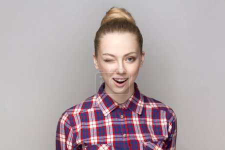 Photo for Portrait of flirting positive adorable woman with bun hairstyle standing blinking her eye, looking at camera, winking, wearing checkered shirt. Indoor studio shot isolated on gray background. - Royalty Free Image