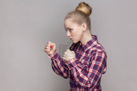 Photo for Side view portrait of angry woman with bun hairstyle standing with clenched fists, being ready to attack somebody, wearing checkered shirt. Indoor studio shot isolated on gray background. - Royalty Free Image