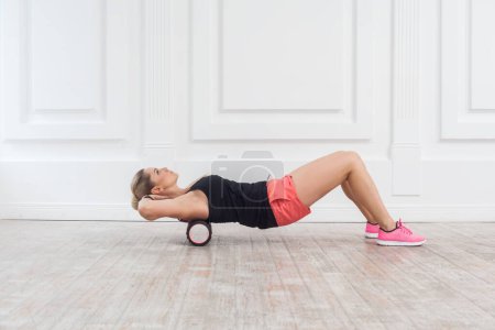 Photo for Side view portrait of slim athletic woman in pink shorts and black top using foam roller in gym to workout to remove the pain, stretching and massaging muscles. Indoor studio shot. - Royalty Free Image