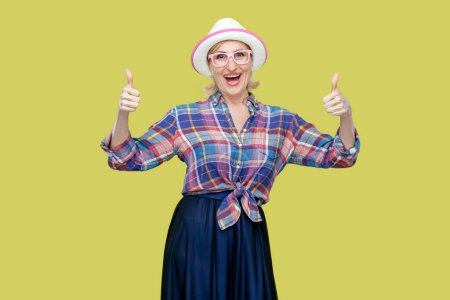 Photo for Portrait of senior woman makes thumbs up sign, demonstrates support and respect to someone, smiles pleasantly at camera, achieves desirable goal. Indoor studio shot isolated on yellow background. - Royalty Free Image