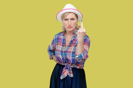 Photo for Portrait of mature woman wearing checkered shirt and hat shows middle finger impolite gesture disrespects someone looks rude at camera. Indoor studio shot isolated on yellow background. - Royalty Free Image