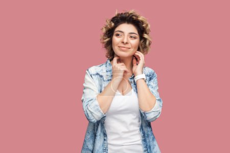 Photo for Portrait of smiling beautiful woman with curly hairstyle wearing blue shirt standing looking away, smiling, dreaming about something pleasant. Indoor studio shot isolated on pink background. - Royalty Free Image