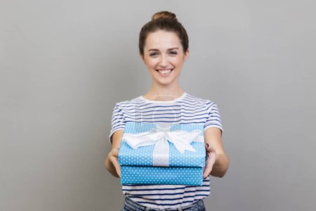 Photo for Portrait of woman wearing striped T-shirt holding out present box, giving gift, looking at camera with optimistic expression, greeting. Indoor studio shot isolated on gray background. - Royalty Free Image